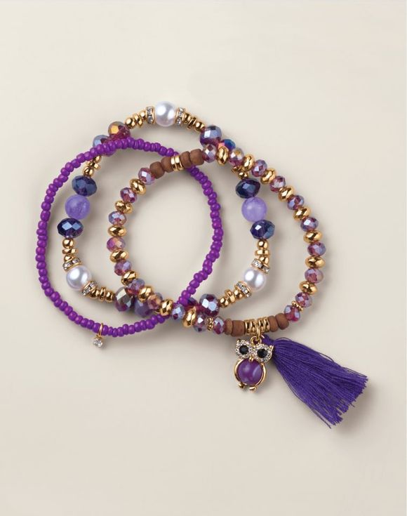 Close-up of a purple and gold beaded bracelet set featuring pearls, crystal accents, wood beads, and an owl charm with a purple tassel. Layered and whimsical with a thoughtful, boho flair.