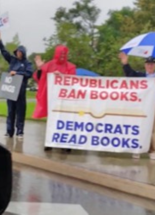 Protesters standing in the rain at the Springfield, Missouri ‘No Kings’ demonstration. One holds a sign reading ‘NO KINGS,’ while another banner reads ‘Republicans Ban Books. Democrats Read Books.’ The group waves at passing cars as rain falls.