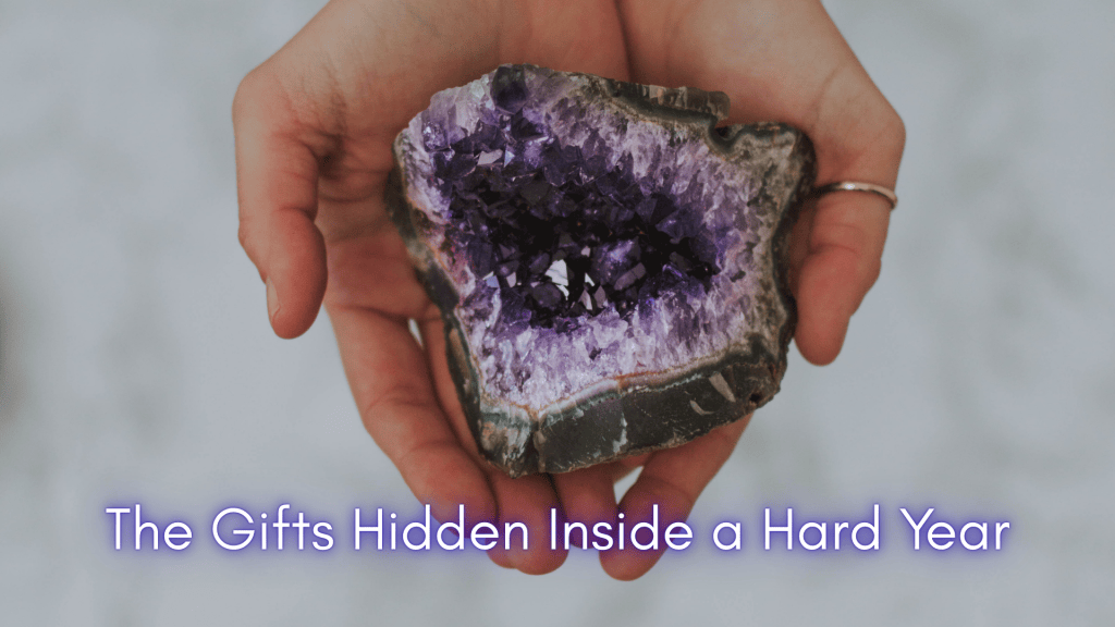 A pair of hands gently holding an open amethyst geode, revealing vibrant purple crystals inside. The image symbolizes the unexpected beauty and inner gifts discovered during a difficult year.