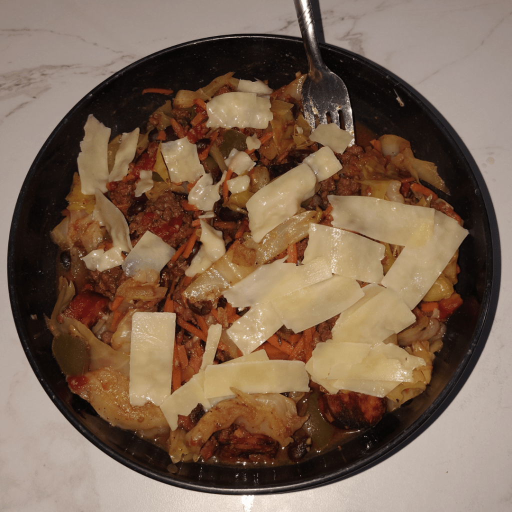 A bowl of dirty cabbage with cabbage, shredded carrots, black beans, ground beef, apple gouda sausage, tomatoes, peppers, and thick Parmesan shreds on top.