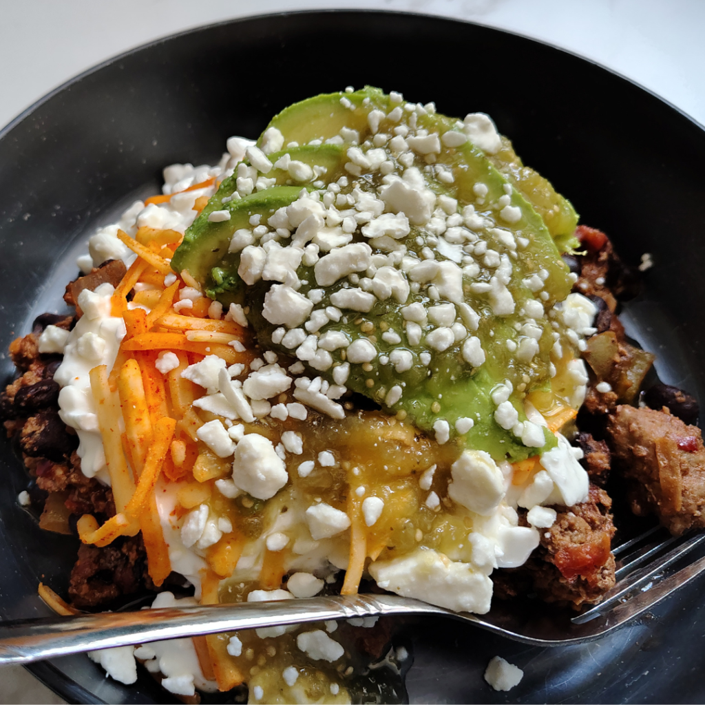Close-up of a high-protein taco bowl topped with cottage cheese, avocado, salsa verde, shredded cheese, and feta over a beef and black bean taco mixture.