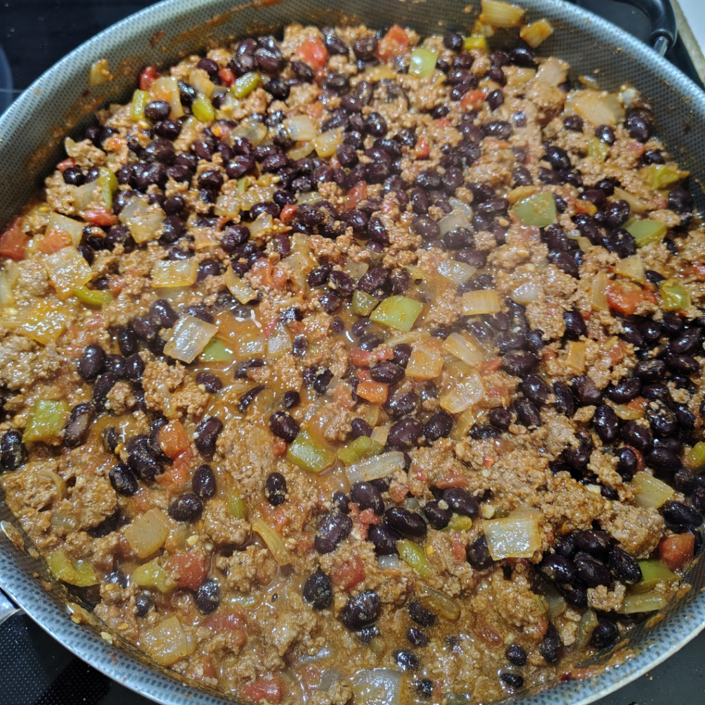 Skillet of taco meat mixture simmering with ground beef, black beans, diced tomatoes, green chilies, onions, green peppers, garlic, and seasoning.
