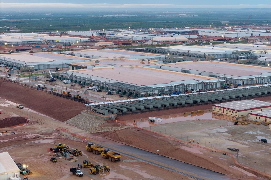 Aerial view of the Stargate AI data center under construction in Abilene, Texas, showing a massive complex of white-roofed buildings, surrounding roads, construction equipment, and rural land.