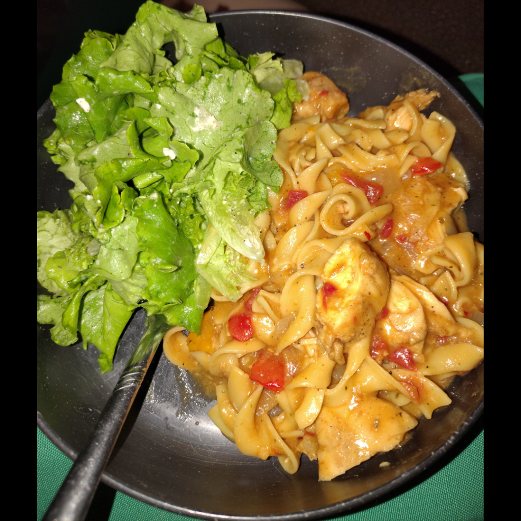 A bowl of Garlic Lover’s Cheddar Parm Chicken Noodles with tender chicken, egg noodles, red peppers, creamy sauce, and a bright lemon-feta salad on the side.