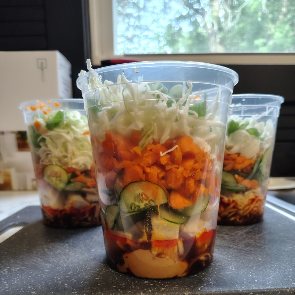 The first heart of palm noodle cup experiment: layered peanut dressing, chicken, cucumbers, carrots, cabbage, and green onions in a 32-ounce container before shaking and serving.