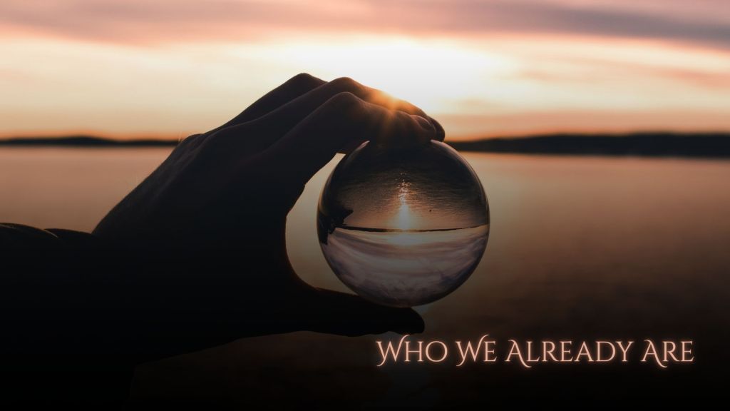 A silhouetted hand holds a clear glass sphere at sunset over calm water, with the horizon and sunlight refracted through the sphere. The title "Who We Already Are" appears in the lower right.
