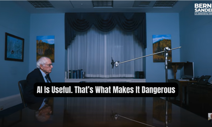 Wide still image from Senator Bernie Sanders’ video showing Sanders seated alone at a long conference table in a dim blue room, facing a large microphone stand across from him. A black rounded text box overlays the lower center with the words: “AI Is Useful. That’s What Makes It Dangerous”.