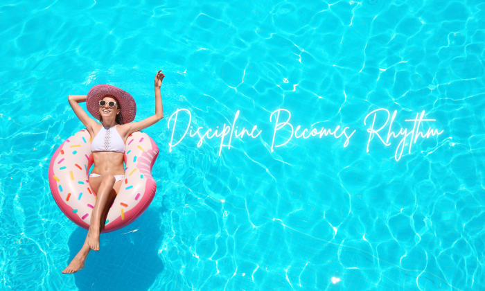 A woman relaxes on a pink pool float in bright blue water with the words “Discipline Becomes Rhythm” overlaid, representing joyful movement, consistency, and building a sustainable fitness routine.