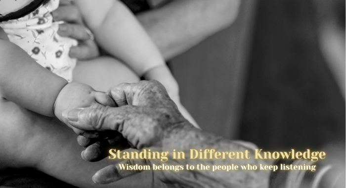 Black-and-white photograph of an elderly hand gently holding the hand of a young child. Gold text reads “Standing in Different Knowledge” and “Wisdom belongs to the people who keep listening.” The image symbolizes learning, connection, and wisdom shared across generations.