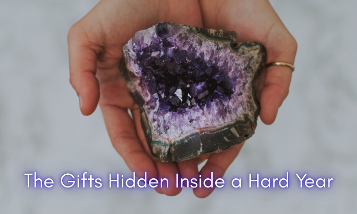 A pair of hands gently holding an open amethyst geode, revealing vibrant purple crystals inside. The image symbolizes the unexpected beauty and inner gifts discovered during a difficult year.